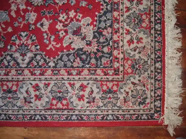 The fringed corner of a red-ground rug with a floral border, on a wooden floor